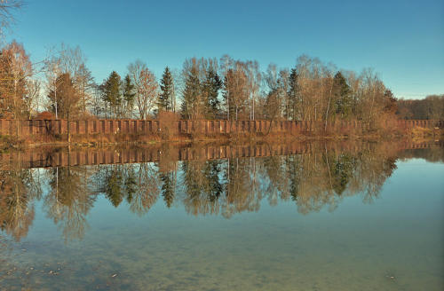 Die Hochwasserschutzwand zwischen Bazhausen und Bayersried spiegelt sich im Balzhauser Baggerweiher.